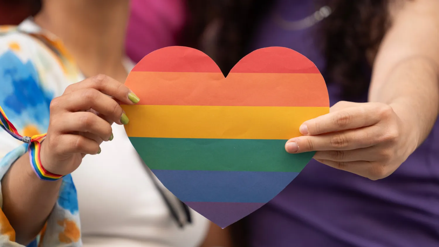 Photo of a heart, which is in pride colors, and is being held by two hands. The people holding the heart are out of focus and out of the frame.