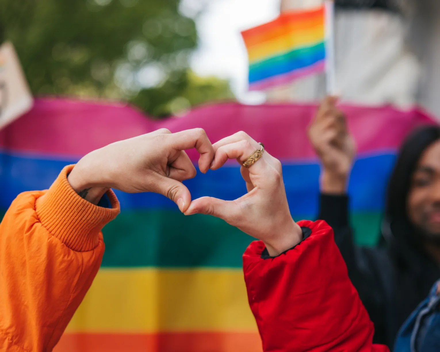 LGBTQ+ flag in the background, with a heart held by hands in the foreground as a symbol of love.