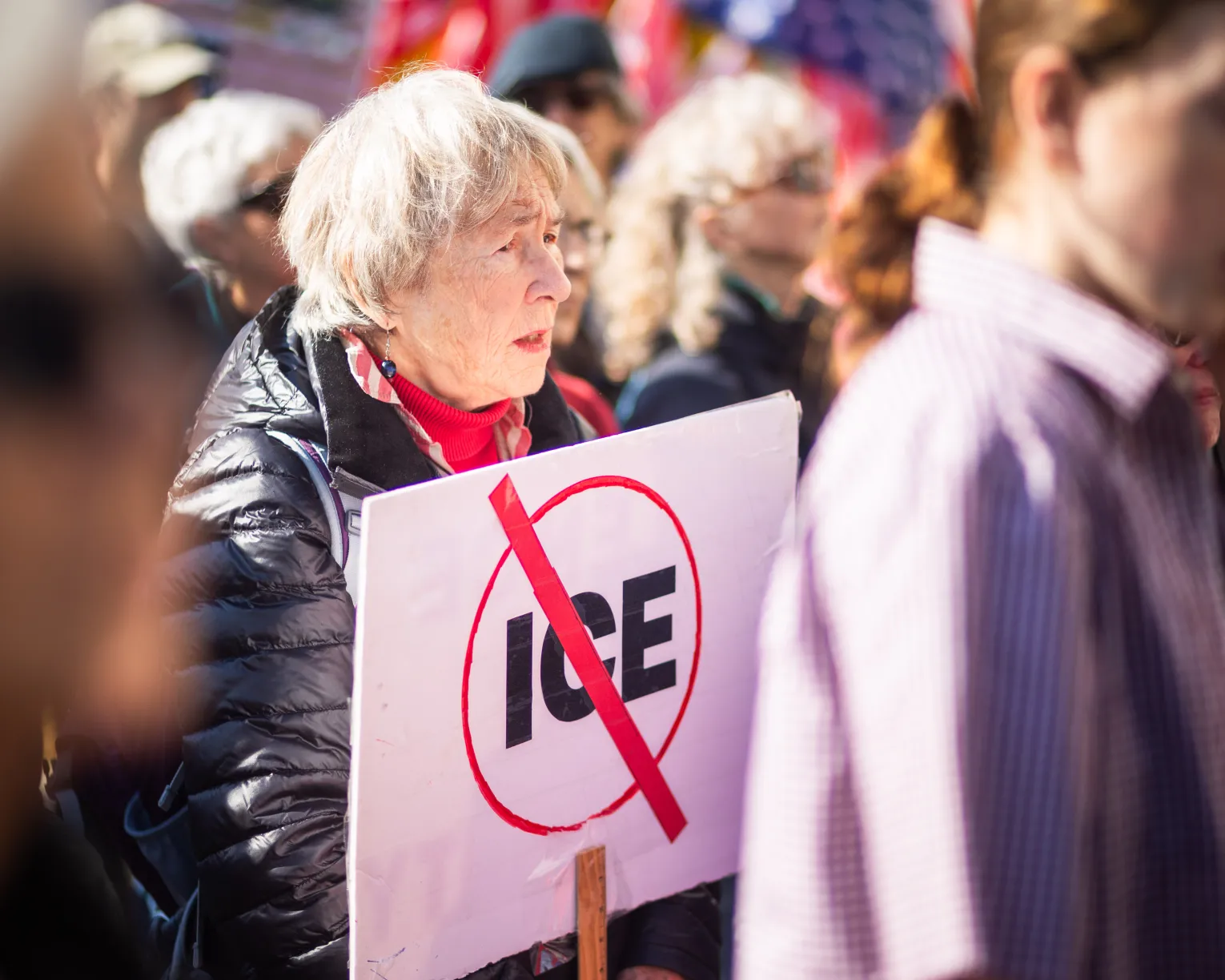 Retired member standing at a rally, holding a sign with a “No ICE” symbol, alongside others advocating for immigrant rights and community protections.