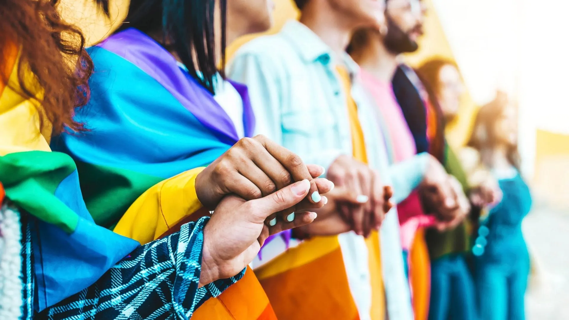 A group of LGBTQIA+ supporters holding hands with a pride flag.