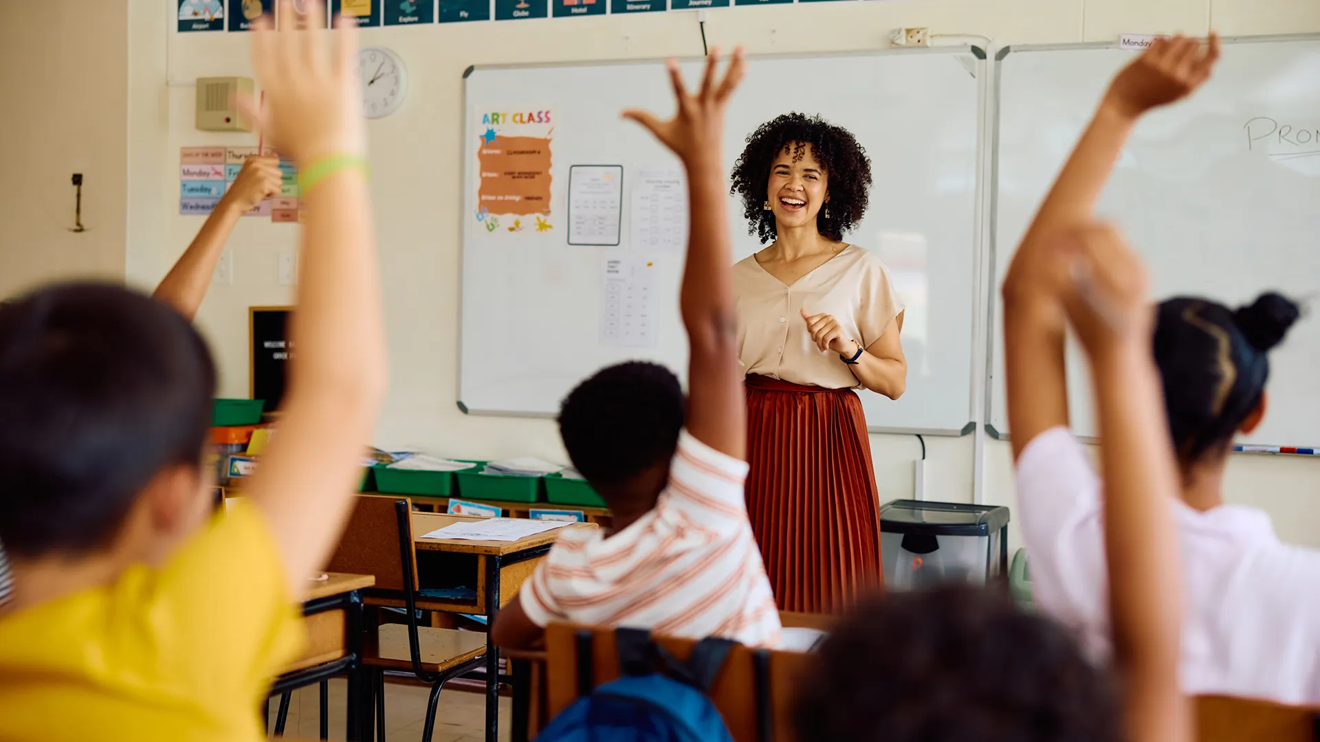 A smiling female teacher standing in the classroom before a handful of students raising their hands. The students are facing the teacher so we can't see their faces.